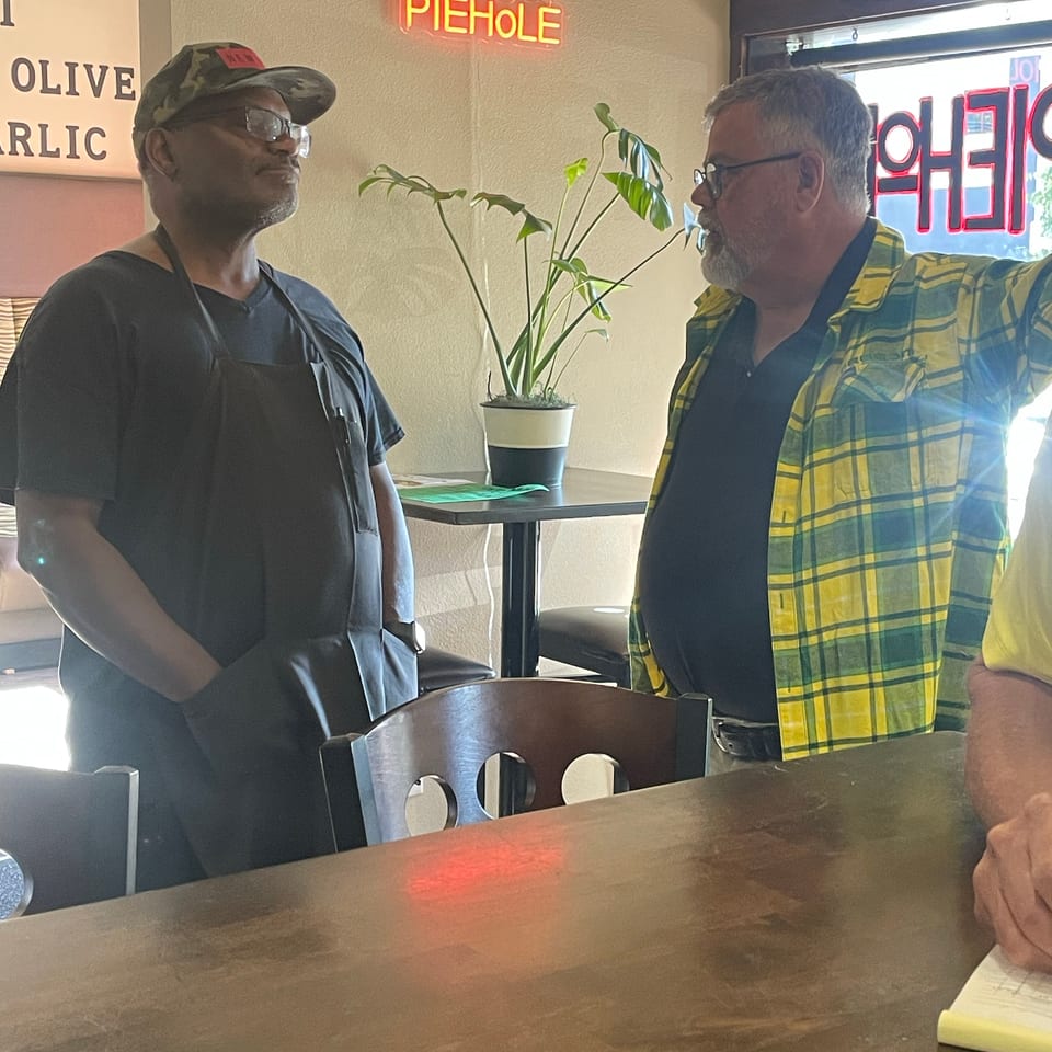 Two men conversing in a restaurant, one in an apron, with a "PIEHOLE" neon sign.
