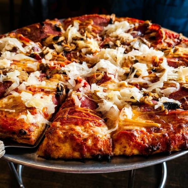 A large, deep-dish pizza with pepperoni, mushrooms, and sauerkraut on a metal stand.