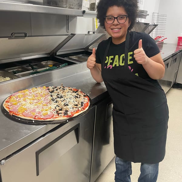 Happy kitchen worker gives thumbs up next to a large half-and-half pizza.