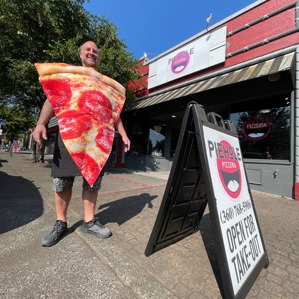 Man in a pizza slice costume stands outside Piehole Pizzeria with an "Open for Take-Out" sign.