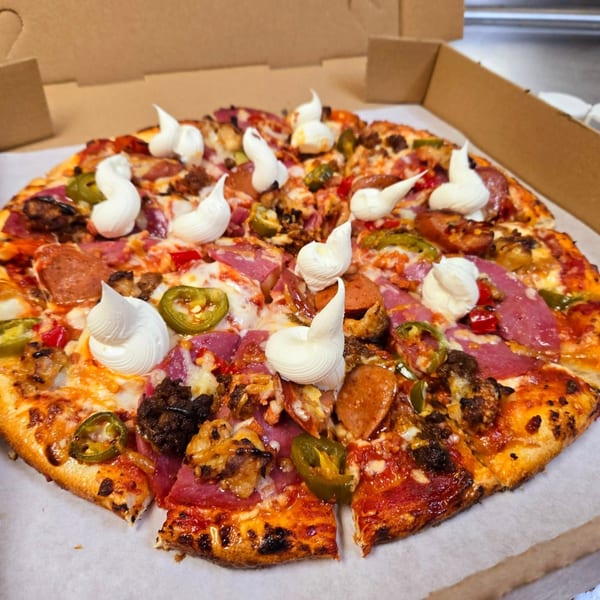 A whole pizza with various meats, jalapeños, and creamy white dollops in a cardboard delivery box.