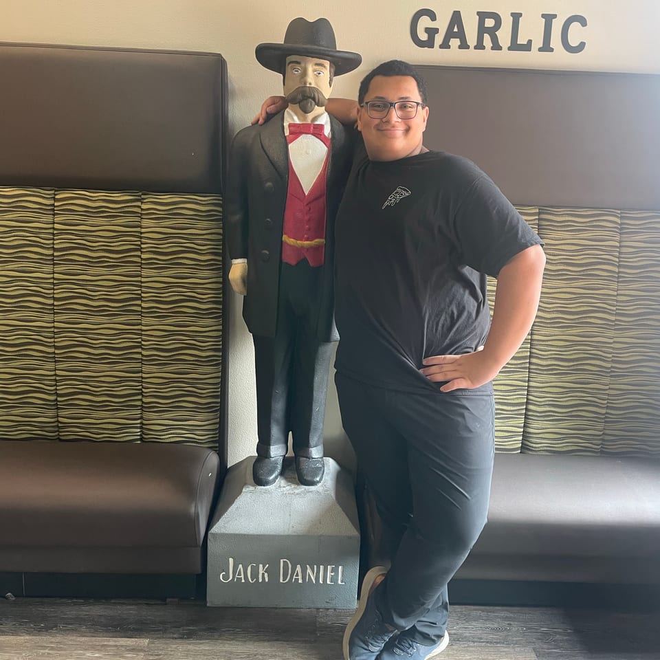 Smiling person with glasses poses next to a Jack Daniel's statue in a restaurant.