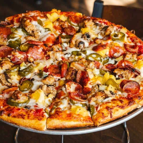 A whole pizza with pepperoni, sausage, pineapple, mushrooms, and jalapeños on a metal stand.