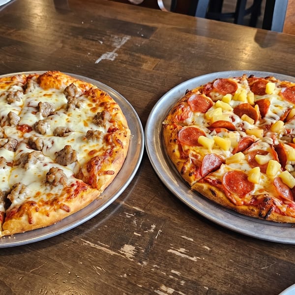 Two pizzas on metal trays, one with sausage and the other with pepperoni and pineapple, on a wooden table.
